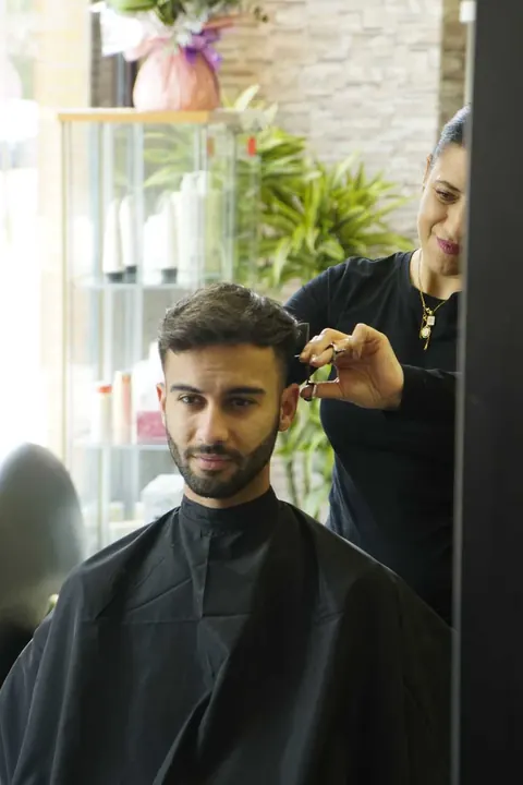 Eva cortando el pelo a tijera a un cliente, vista a través del espejo